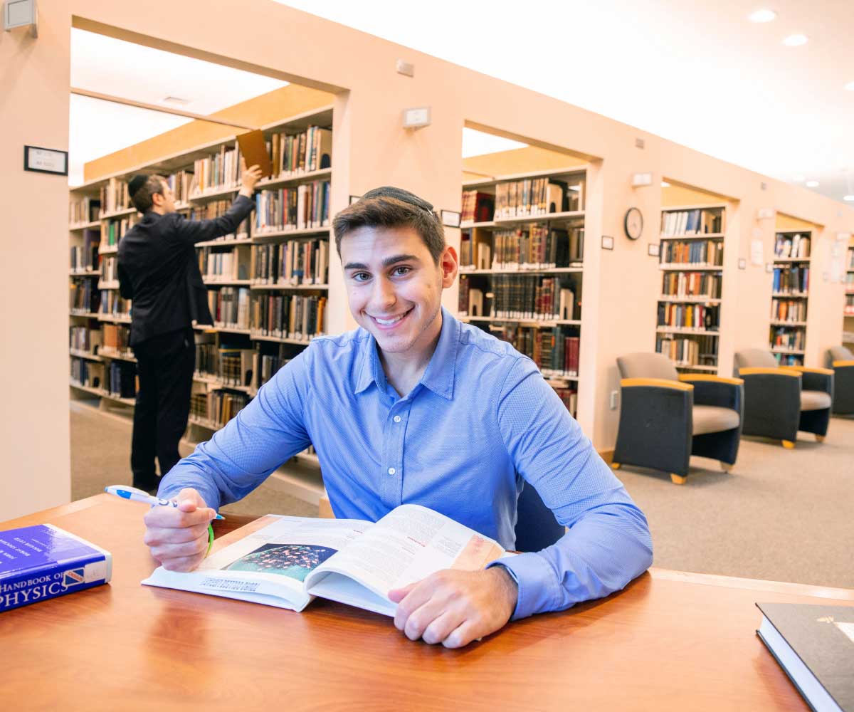 Happy student reading a textbook in the library, with many bookshelves in the background